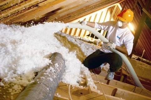Technician in yellow hardhat installing white blown-in insulation in an attic.