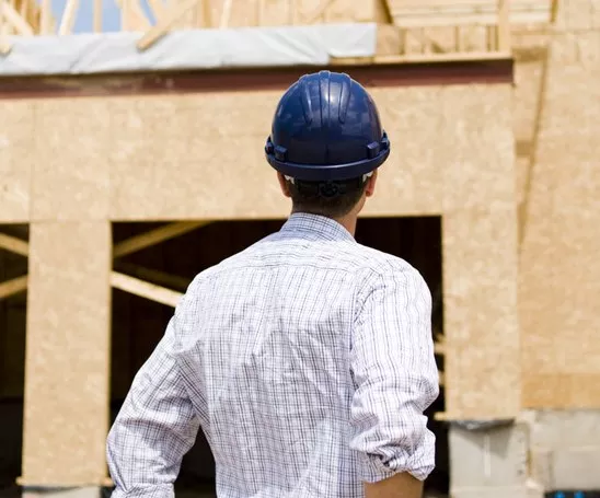 A contractor turned away from the camera, wearing button-down shirt and blue hard hat, looking at in-construction building.