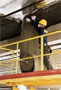 Technician in yellow hard hard installing batt insulation on ceiling of a commercial building.